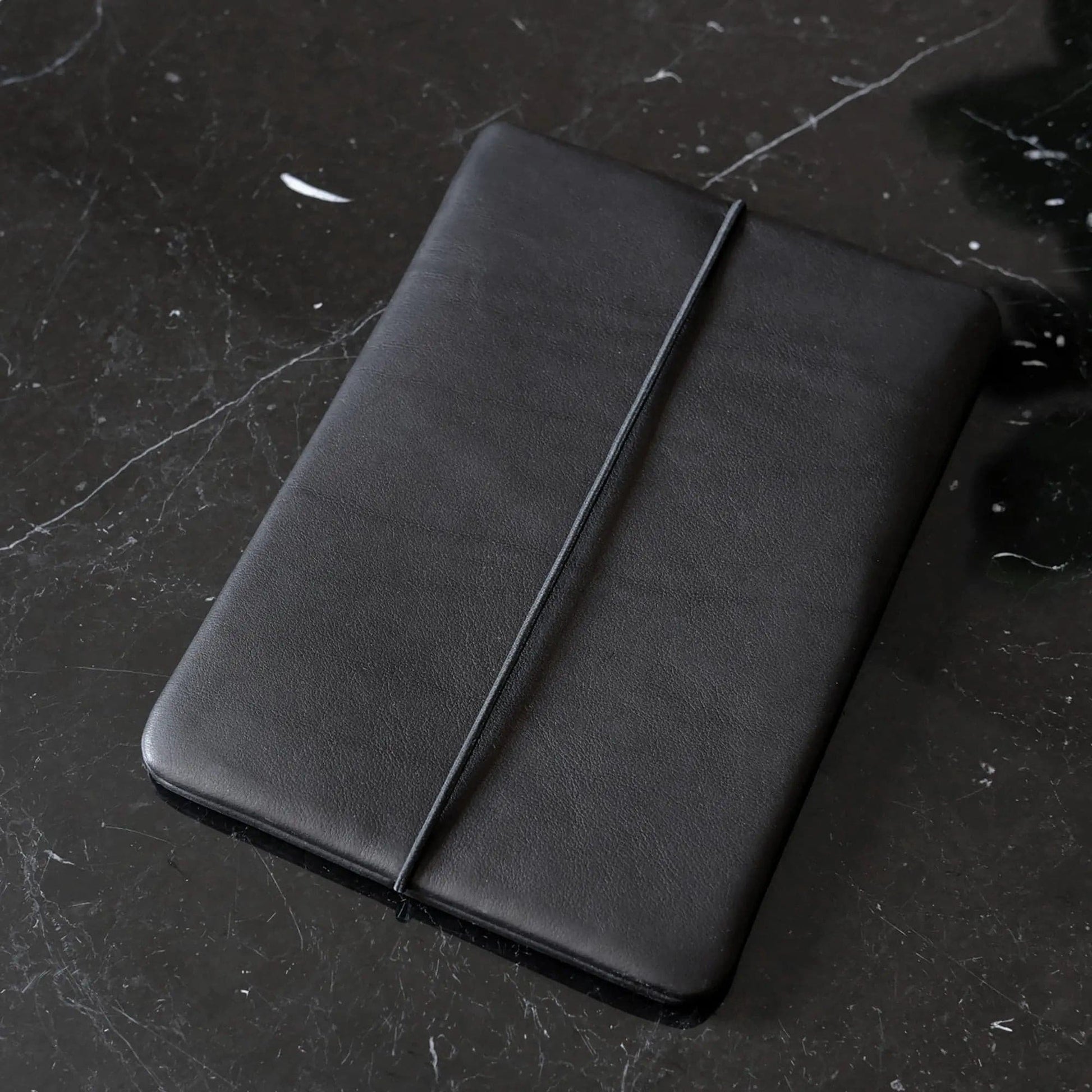 Macbookhülle aus schwarzem Leder liegt schräg auf einem schwarzem Marmortisch