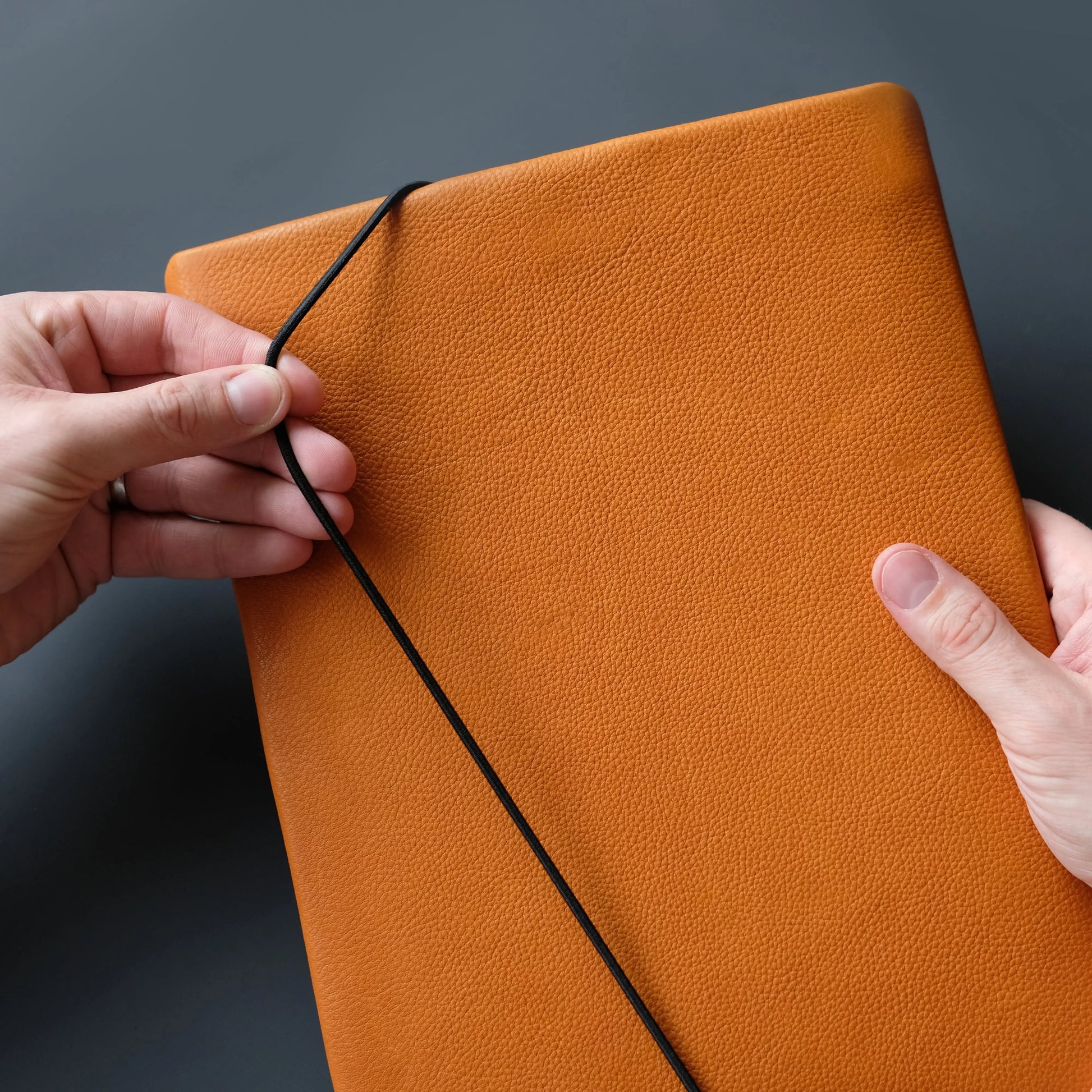 Macbookhülle aus orangefarbenem Leder wird mit schwarzer Gummikordel von zwei Händen geschlossen