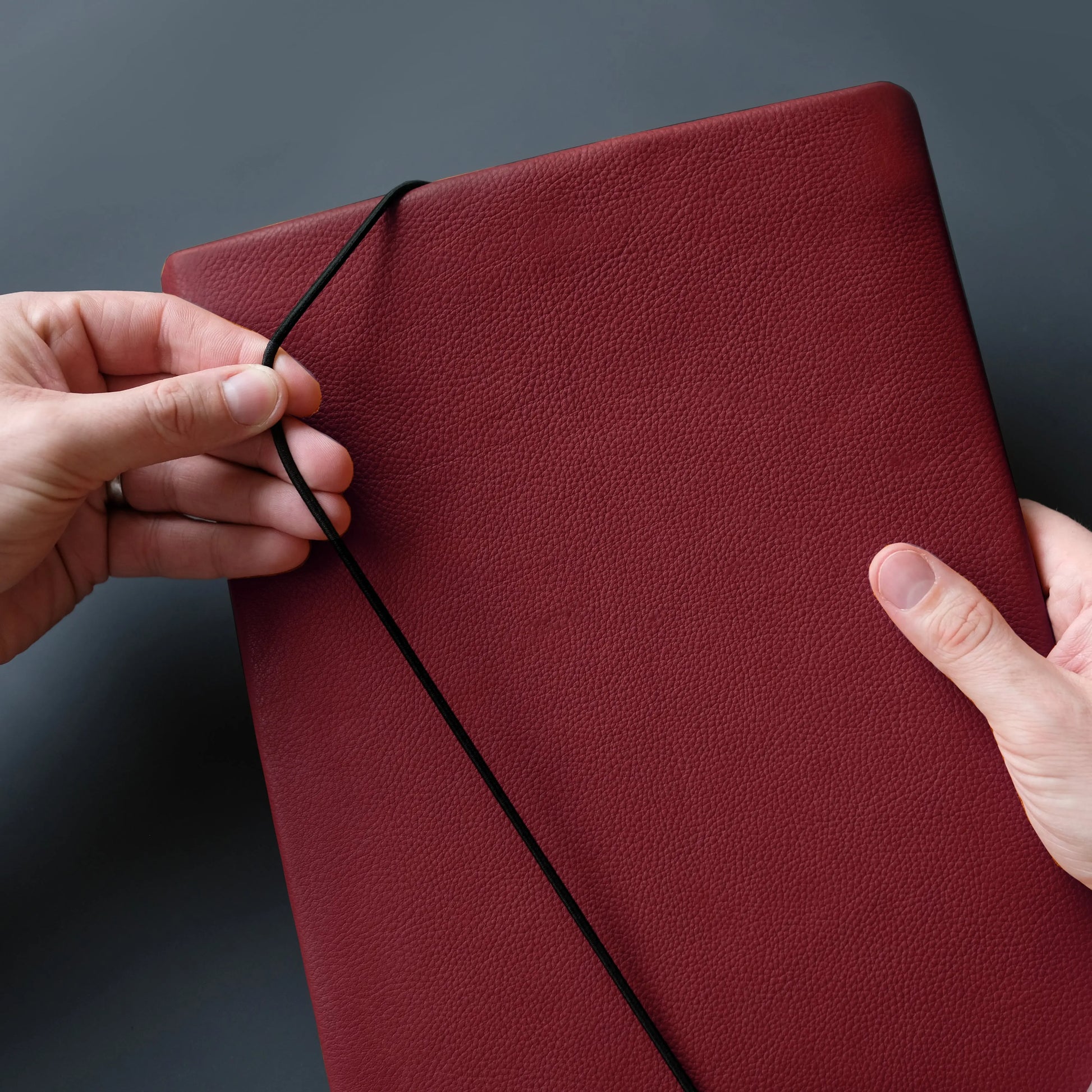 iPadhülle aus weinrotem Nappaleder, die von zwei Händen mit der Gummikordel verschlossen wird.