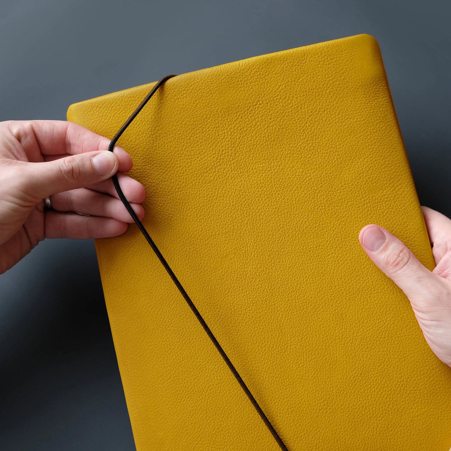 Hülle aus Leder für Tablets in gelb mit schwarzem Gummi das von einer Hand geschlossen wird.